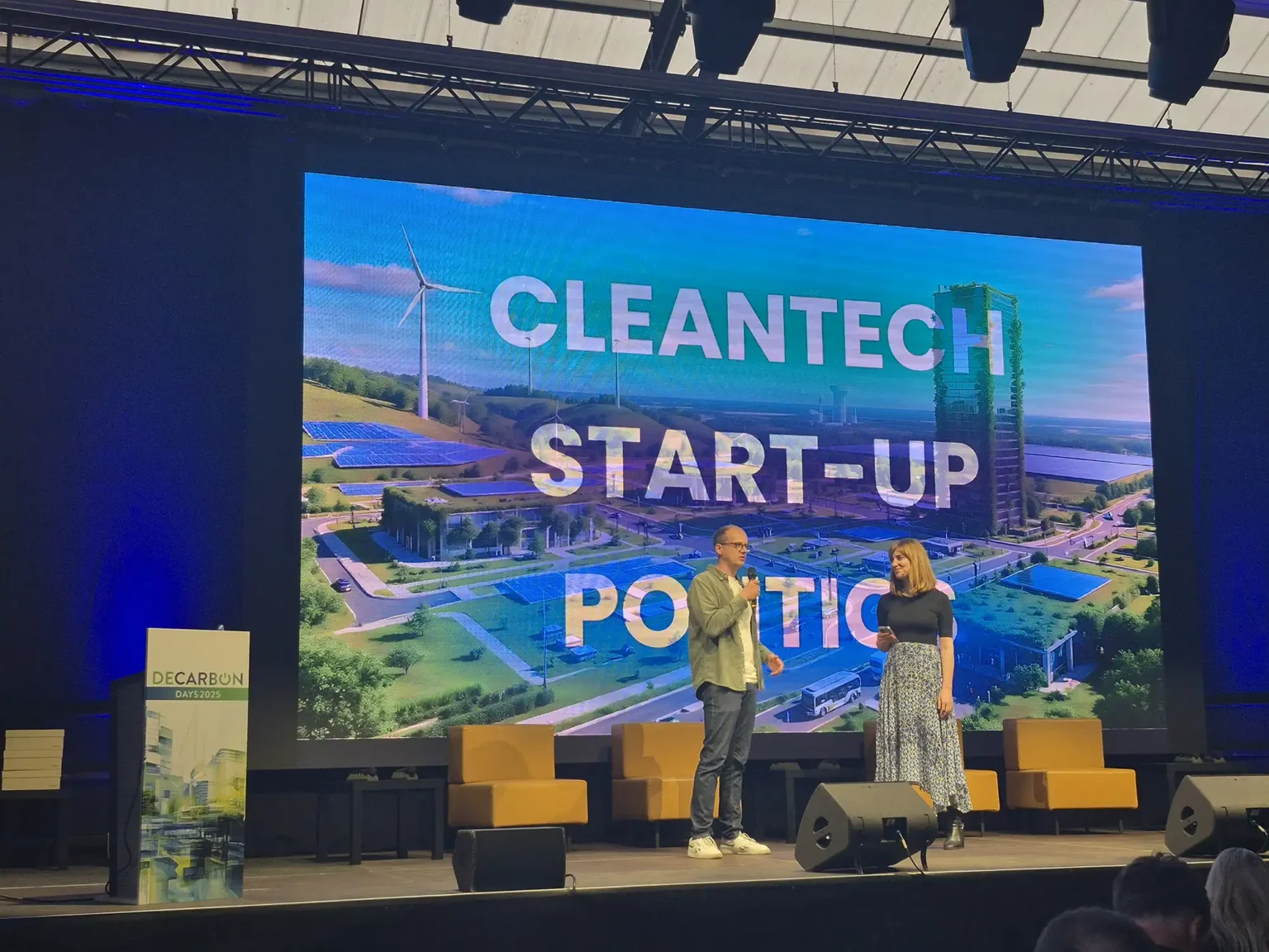 Zu sehen ist eine Bühne, auf der zwei menschen stehen, dahinter ein Banner mit dem Text "Cleantech Start-Up Politics"